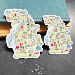 May include: Two stickers in the shape of the state of Michigan, featuring a colorful illustrated map with landmarks and city names. The stickers are on a dark surface with a book in the background.