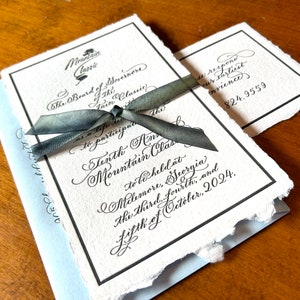 May include: A white invitation with black calligraphy text announcing the Tenth Annual Mountain Classic to be held in Melemore, Georgia on October 3rd, 4th, and 5th, 2024. The invitation is tied with a gray ribbon and has a small RSVP card attached.
