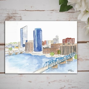 Puede incluir: Pintura acuarela de un horizonte urbano con edificios altos y un puente azul sobre un río. Los edificios son en tonos de marrón, gris y azul. El puente es un puente de armadura de acero con una barandilla azul. El río es de color azul claro. La pintura está firmada "Anthony J. Davis 2018".