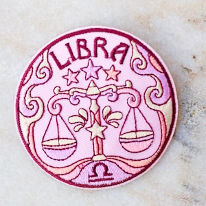 May include: A round, embroidered patch featuring the Libra zodiac symbol. The patch is primarily pink with a dark pink border and accents. The word "LIBRA" is arched across the top, with stars and a scale design in the center.