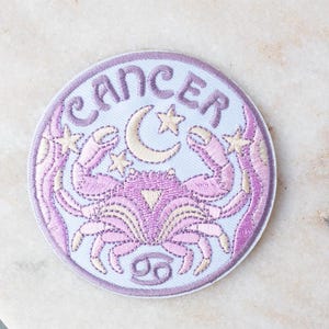 Op de afbeelding: Geborduurde patch met het sterrenbeeld Kreeft. Het ontwerp bevat een krab, een maan, sterren en het woord "CANCER" in lichtpaars en crème. De patch is rond en heeft een lichtblauwe achtergrond.