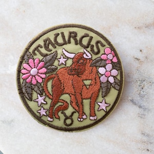 May include: Round embroidered patch featuring the Taurus zodiac symbol. The patch has a light green background with the word "TAURUS" in a dark green font. The design includes a brown bull, pink flowers, and stars.