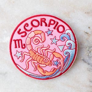 Op de afbeelding: Geborduurde patch met het woord "SCORPIO" in rood, rond een roze cirkel. De patch bevat een schorpioen, sterren en golven in tinten oranje, roze en blauw. Het Schorpioen-symbool is ook aanwezig.