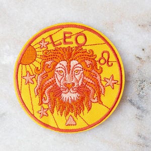 以下が含まれることがあります： オレンジと赤色のライオンの頭が描かれた、丸い黄色の刺繍パッチ。「LEO」の文字が上部にあり、星と太陽のグラフィックがあります。パッチには赤い縁取りがあります。