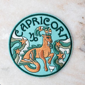 Op de afbeelding: Een ronde geborduurde patch met het woord "CAPRICORN" in groene letters. De patch heeft een lichtblauwe achtergrond met een illustratie van een geit-vis hybride, sterren en golfachtige ontwerpen in groen, oranje en wit. Ongeveer 9 cm in diameter.