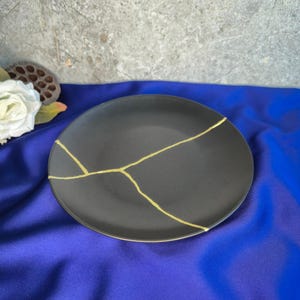 May include: A black ceramic plate with gold lines, demonstrating the Japanese art of Kintsugi. The round plate is displayed on a blue surface. The gold lines highlight the repaired cracks, adding a unique aesthetic to the dish.