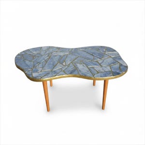 May include: A coffee table with an irregular, kidney-shaped top featuring a blue and grey stone mosaic pattern with gold-toned grout lines. The table has a gold-toned edge and four tapered, light brown wooden legs extending downward.
