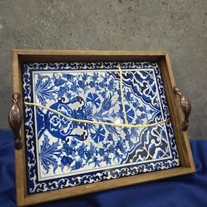 May include: Rectangular wooden serving tray with a blue and white floral tile insert. The tray has bronze-colored handles and a light brown wooden frame. The tile features a detailed floral pattern with a white background.