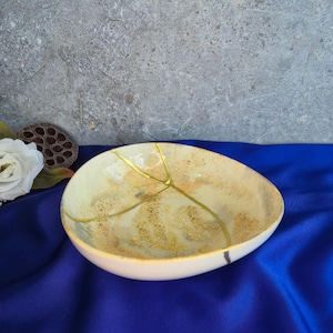 May include: A cream-colored, oval-shaped ceramic bowl with gold-colored kintsugi-style repair lines. The interior has a mottled beige and brown pattern. The bowl sits on a blue surface, with a white rose and seed pod in the background.