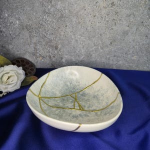 May include: White and blue ceramic bowl with gold-colored kintsugi-style repairs. The bowl has an oval shape and a speckled blue and white interior. The gold lines create a unique pattern across the surface.