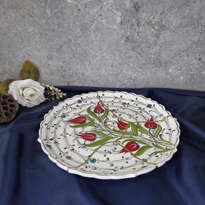 May include: A white ceramic plate with a decorative design of red tulips and green leaves. The plate is cracked and repaired with gold-colored epoxy.