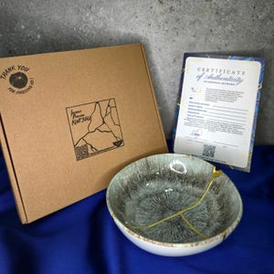 Può includere: Una ciotola in ceramica Kintsugi con linee di riparazione dorate, una scatola di cartone marrone con un logo e un certificato di autenticità. La ciotola ha un bordo bianco e un interno grigio e blu. La scatola ha la scritta "Thank you for choosing us."