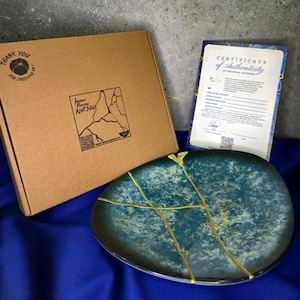 May include: A teal and gold Kintsugi plate with a certificate of authenticity and a brown box. The plate has gold-colored lines that repair the cracks. The box has a logo and the text "Thank you for choosing us!"