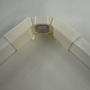 May include: A white, angular corner piece with a clear, oval-shaped insert. The piece is composed of several rectangular sections joined at right angles, creating a sturdy, geometric form. The design suggests a structural or architectural component.
