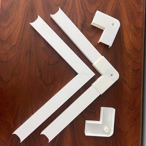 Puede incluir: Piezas de esquina y tiras de conexión de plástico blanco dispuestas sobre una superficie de madera oscura. Las piezas parecen ser componentes para un proyecto de enmarcado o construcción similar. Los componentes son nuevos.