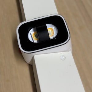 May include: A white rectangular lighting fixture featuring a central black square housing with a rounded frame. Inside the black housing, a horizontal light strip with two visible yellow LED components is bisected by a small black vertical clip.