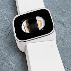 May include: A white rectangular device with a black central display area featuring a small horizontal black strap over a white interior with two yellow light components. The device has a long white band extending from both ends.
