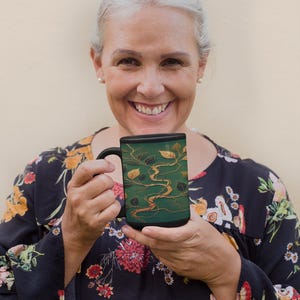 May include: A woman is holding a black and green mug with a gold design of a river and leaves. The mug is tall and has a handle.