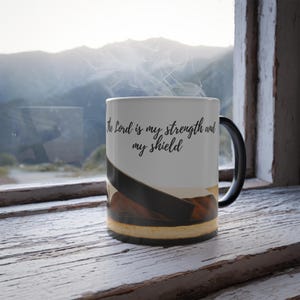 May include: White ceramic mug with a black handle. The mug has a black and brown image of a book on the bottom half. The top half of the mug has the text "The Lord is my strength and my shield" in black.