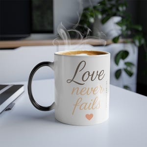 May include: White ceramic coffee mug with a black handle. The mug has the text "Love never fails" in brown lettering with a small pink heart at the bottom. The mug is filled with coffee and steam is rising from the top.