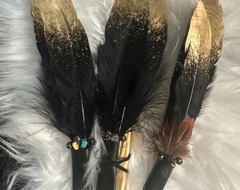 May include: Three decorative feathers with black and gold colouring. The feathers have a black base with gold tips. Each feather is attached to a black leather handle with small beads and accents. The feathers are displayed on a white, fluffy surface.
