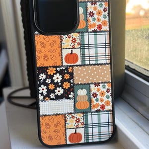 May include: Black phone case with a patchwork design featuring pumpkins, flowers, and fall leaves. The case has a black border and a camera cutout. The design includes orange, white, and brown colors.