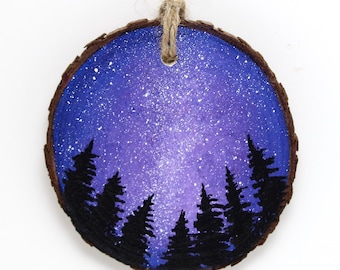 May include: A round, wooden ornament with a painted night sky scene. The background is a gradient of purple and blue with white speckles. Silhouetted black pine trees are at the bottom. A natural twine loop is attached for hanging.