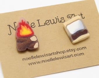 May include: A pair of polymer clay earrings featuring a campfire and a s'more. The campfire is brown with orange flames. The s'more is white with brown chocolate and a yellow graham cracker.