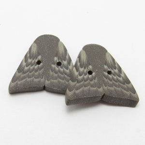 Puede incluir: Dos botones grises en forma de ala con patrones de plumas detallados. Cada botón tiene dos agujeros para coser. Los botones están hechos de un material moteado y mate, sobre un fondo blanco.