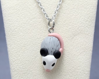 May include: A silver-toned metal chain necklace featuring a sculpted opossum pendant. The opossum has a white face, black ears, a gray textured body, and a light pink tail, all crafted with a smooth, matte finish.