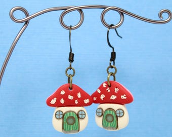 May include: A pair of whimsical mushroom house earrings. Each earring features a red cap with white spots, a white base with a green door, and small blue window details. The earrings hang from dark metal hooks.