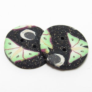 Op de afbeelding: Twee ronde knopen met een zwarte achtergrond besprenkeld met gouden glitter. Elke knoop heeft een ontwerp van een lichtgroene mot met paarse accenten en een witte halve maan. De knopen hebben twee gaten om op te naaien.