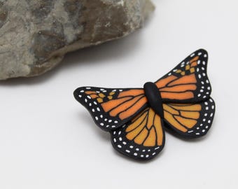 May include: A detailed, handcrafted butterfly pin with orange and black wings, accented with white dots. The butterfly has a black body and is set against a white background, with a gray stone in the upper left corner.