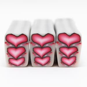 May include: Three rectangular polymer clay beads with a white base and a repeating pattern of three stacked pink hearts. The hearts have a dark pink outline and a lighter pink interior. Small pink dots accent the design.