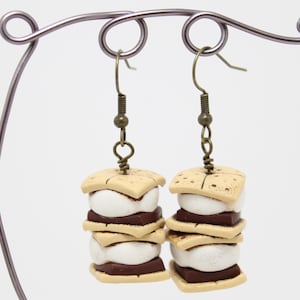 May include: A pair of dangle earrings featuring a stacked s'more design. Each earring has three layers of graham crackers, chocolate, and marshmallow. The earrings are made of polymer clay and have bronze-colored hooks.