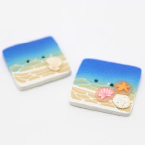 May include: Two square-shaped decorative tiles with a beach theme. Each tile features a blue sky, sandy beach, and seashells. One tile has a single shell, while the other has a starfish and multiple shells.