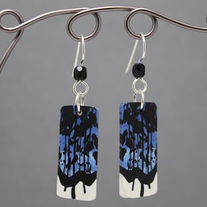 Puede incluir: Un par de pendientes rectangulares con un diseño abstracto en negro y azul. Los pendientes tienen una base blanca con gotas negras. Cada pendiente está suspendido de un gancho plateado con una cuenta negra.