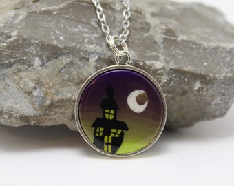 May include: Silver-toned pendant necklace featuring a round charm with a spooky house silhouette under a crescent moon. The charm has a purple and yellow gradient background. The necklace is a Halloween-themed accessory.