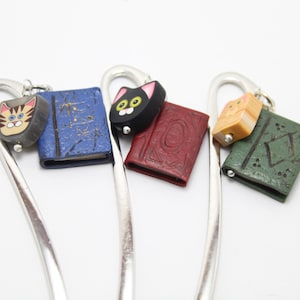 Puede incluir: Un conjunto de marcadores de metal plateado, cada uno adornado con un dije. Los dijes incluyen una cara de gato, un libro azul, una cara de gato negro, un libro rojo, un libro beige y un libro verde. Fondo blanco.