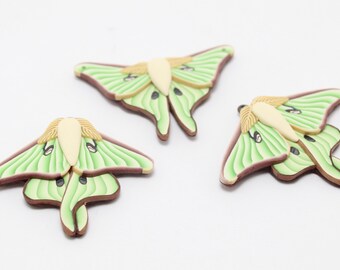 May include: Three handmade moth-shaped polymer clay ornaments. Each moth features light green wings with brown edges and a cream-colored body. The wings have detailed vein patterns. These decorative ornaments are ideal for crafting or home decor.