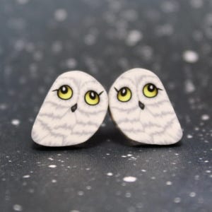 May include: A pair of owl-shaped earrings. Each earring features a white owl with gray markings, yellow eyes, and a small black beak. The earrings are set against a dark background with white speckles.