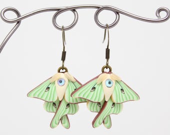 May include: A pair of handmade earrings featuring a luna moth design. The earrings have a light green and brown color scheme, with a small blue gem in the center of each moth. The earrings are suspended from a metal display stand.