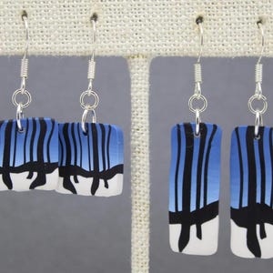 Puede incluir: Dos pares de pendientes rectangulares con un diseño degradado azul y blanco, acentuados por líneas negras verticales. Los pendientes están suspendidos de ganchos plateados, mostrando un estilo artístico único.