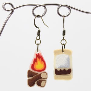 Puede incluir: Un par de pendientes colgantes con un diseño de fogata y un s'more. El pendiente de fogata es marrón y blanco con una llama roja y amarilla. El pendiente de s'more es beige con un malvavisco blanco y chocolate marrón.