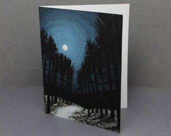 May include: Greeting card featuring a nighttime forest scene. The artwork depicts tall, dark trees lining a path under a starry sky and a full moon. The card is white on the inside and has a matte finish.