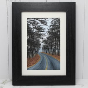 May include: Framed landscape painting of a winding road through a forest. The road is gray with yellow lines, flanked by tall, dark trees. The artwork is in a black frame with a white mat, creating a serene, natural scene.