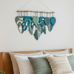 May include: A bedroom with a wooden bed frame, white bedding, and green and beige cushions. A fringed, woven throw is at the foot of the bed. Above the bed hangs a wall decoration of teal and white leaf shapes. The bed is approximately 150cm wide.