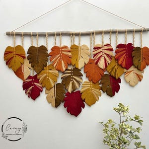 May include: A wall hanging made of macrame with a leaf design. The leaves are in shades of red, orange, yellow, and brown. The leaves are hanging from a wooden dowel rod.