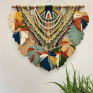 Puede incluir: Un tapiz de macramé de pared colorido con un diseño geométrico y formas de hojas. El tapiz está hecho con una variedad de fibras naturales y presenta cuentas de madera.