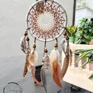 May include: A white and brown dreamcatcher with feathers and wooden beads. The dreamcatcher has a circular frame with a web-like pattern and a smaller circle in the centre. The feathers are brown, white, and orange.
